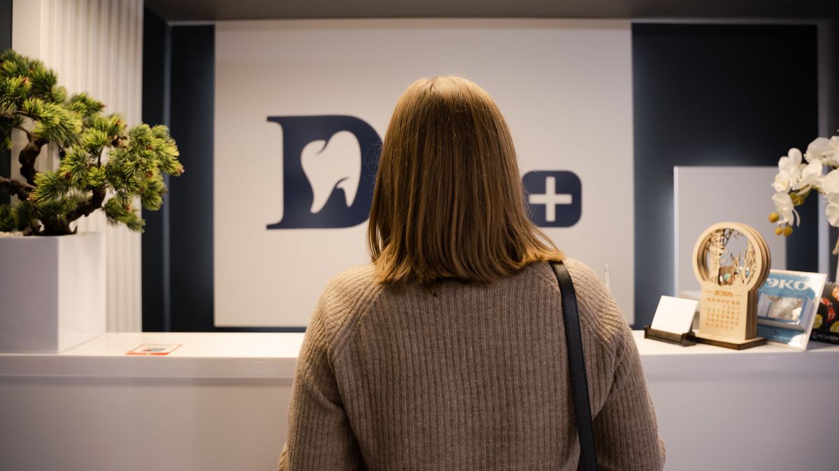 A woman stands at the dental clinic reception wearing a brown sweater, facing away.