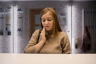 A woman in a brown sweater experiencing a toothache, holding her cheek indoors.