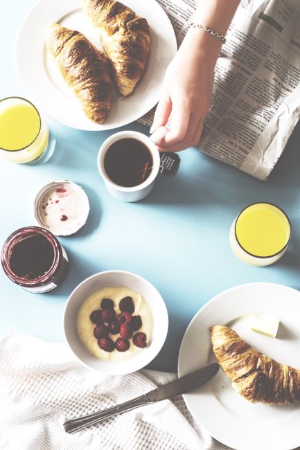 A hotel breakfast table with sliced baguette, croissants, butter and jam, a bowl of mixed fruit, yogurt cups, and small glasses of orange juice next to a coffee pot.
