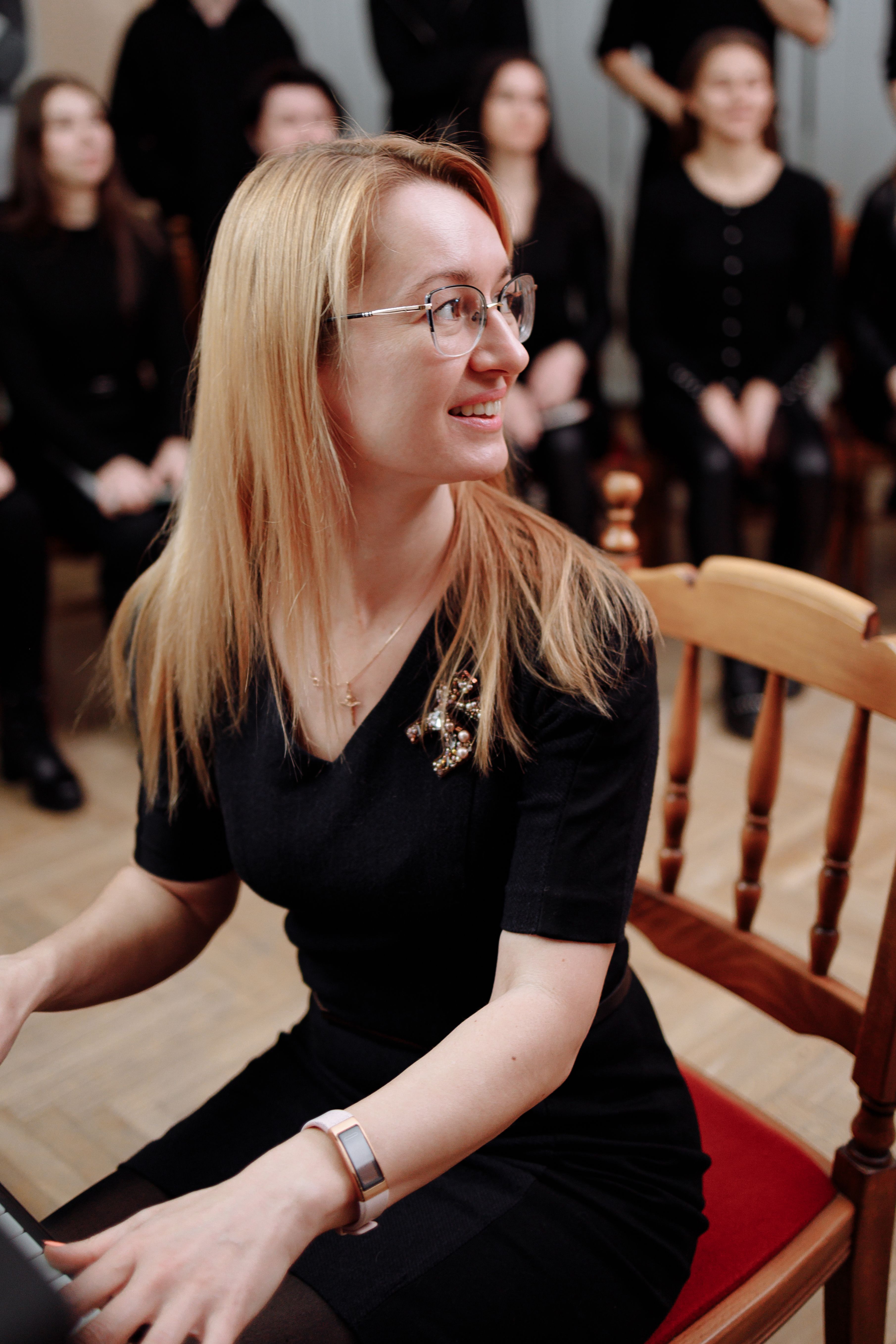 Blond woman in a black dress with glasses, smiling while seated indoors.