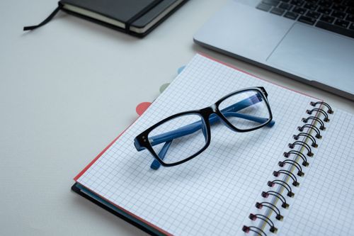 Free Close-up of black eyeglasses resting on an open graph paper notebook near a laptop. Stock Photo