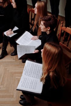 Group of women in a choir holding music sheets, seated on wooden chairs, practicing indoors.