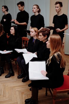 Group of choral singers holding sheet music during a rehearsal in a room.