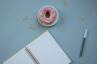 Donut on a Plate Near a Spiral Notebook and Paper Clips