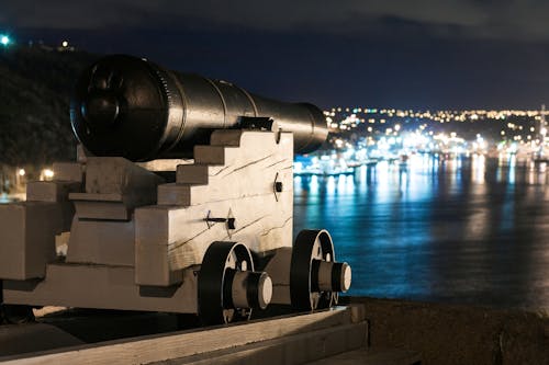 Vintage artillery cannon on wooden support on waterfront of city with glowing lights at night