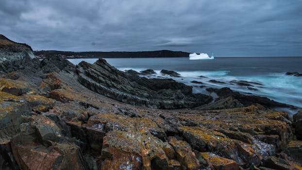 A stunning seascape showcasing rocky shoreline and an iceberg under overcast skies, capturing nature's power.