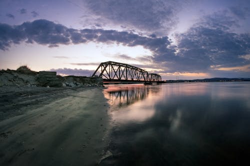 Metal bridge over reflecting river with sandy beach under cloudy sky at sundown