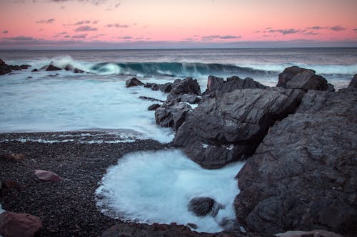 Free Breathtaking view of wavy ocean with foamy flows against beach with rocky formations under cloudy sky at sunset Stock Photo