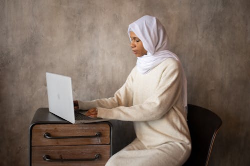 Side view of ethnic female in headscarf typing on netbook placed on wooden cabinet