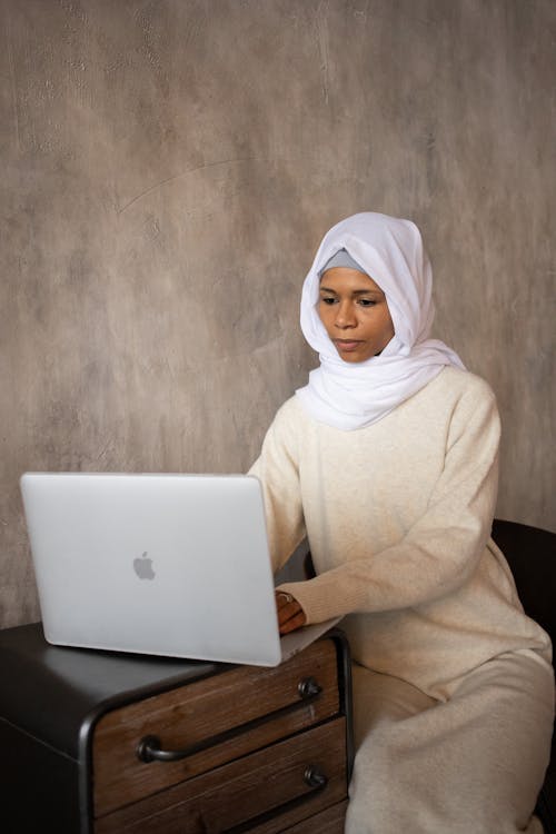 Young ethnic pensive Muslim female in hijab typing report on netbook placed on timber cabinet