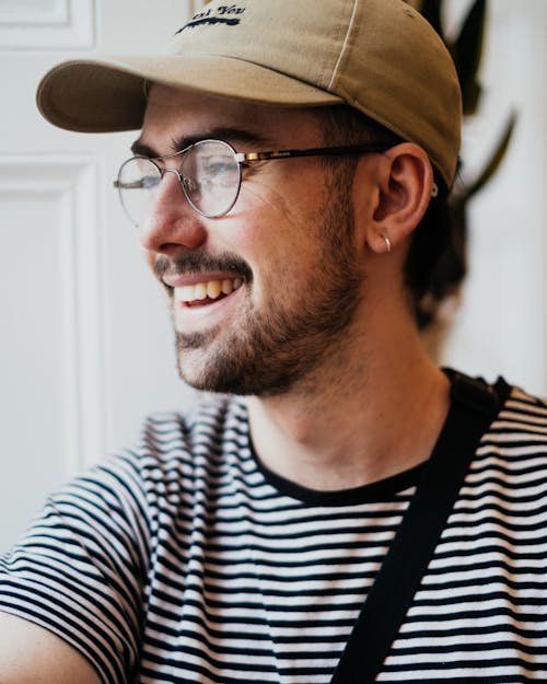 Happy unshaven young guy in casual cap and eyeglasses smiling and looking away in light room