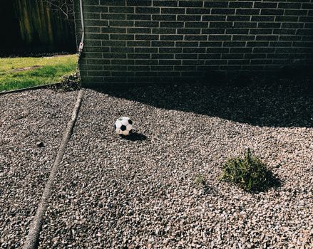 High angle of pebble ground with football ball near small plant and building with brick wall near green grassy lawn and fence in backyard in sunny summer day