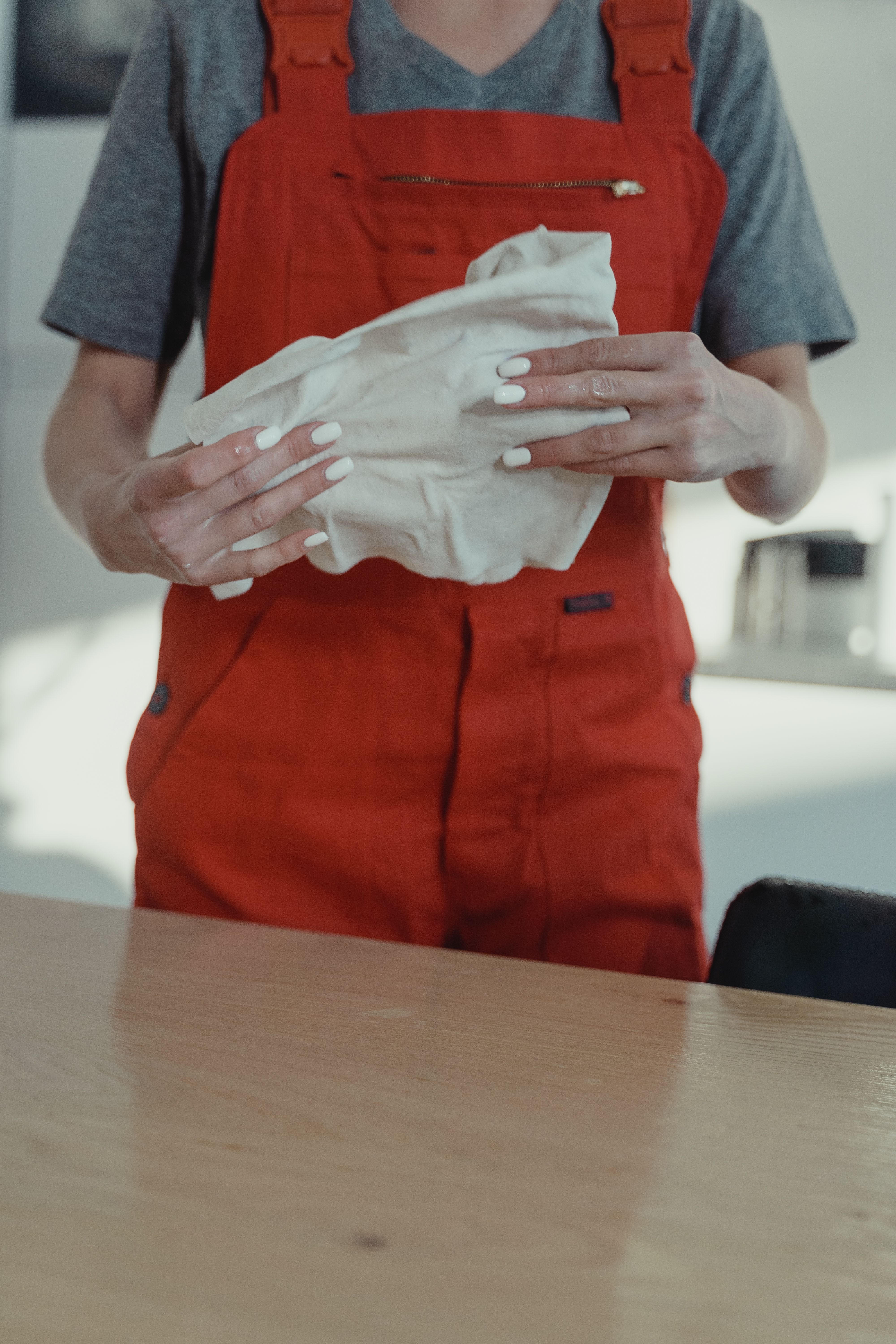 A woman wearing red overalls cleans a wooden table with a white cloth indoors. - Daily Care Routines for Handbags