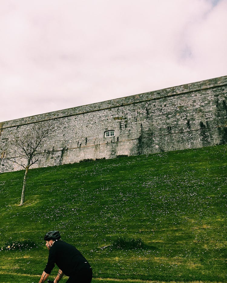 Faceless Man In Helmet Near Grassy Field With Stone Fence