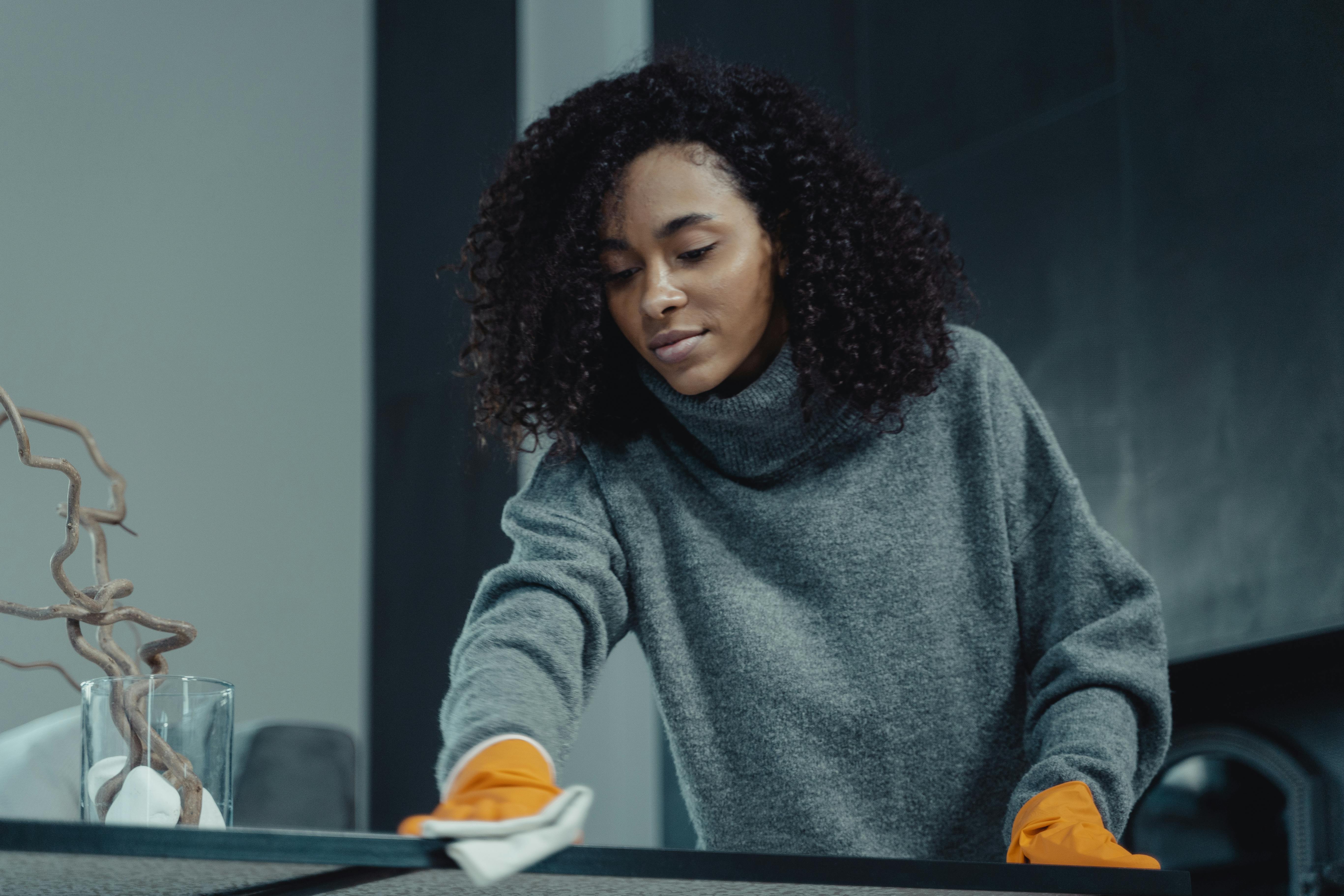 A woman in a gray sweater wipes a table with care, showcasing home cleaning in comfort.
