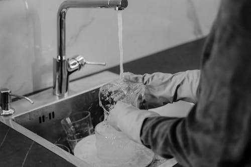 Grayscale Photo of a Person Washing a Glass in the Kitchen Sink
