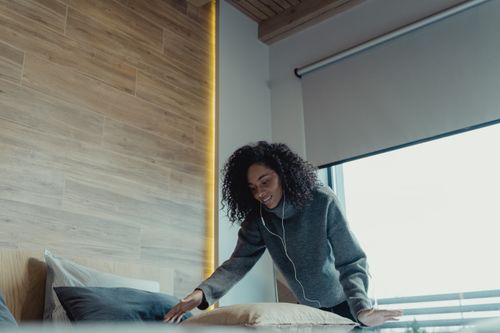 Free Smiling woman with curly hair arranging pillows while listening to music indoors. Cozy indoor setting. Stock Photo