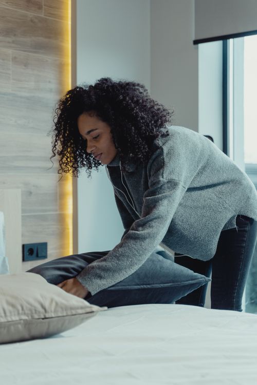Free A woman making a bed in a modern bedroom with natural light, reflecting a cozy and tidy home environment. Stock Photo