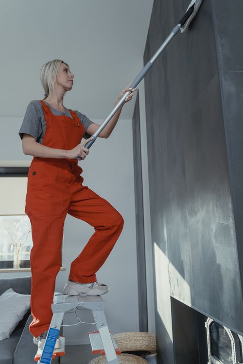 Free Woman in red overalls cleaning a wall using a mop while on a ladder indoors. Stock Photo