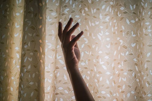 Crop delicate hand of unrecognizable female near window against rustic curtains in daylight