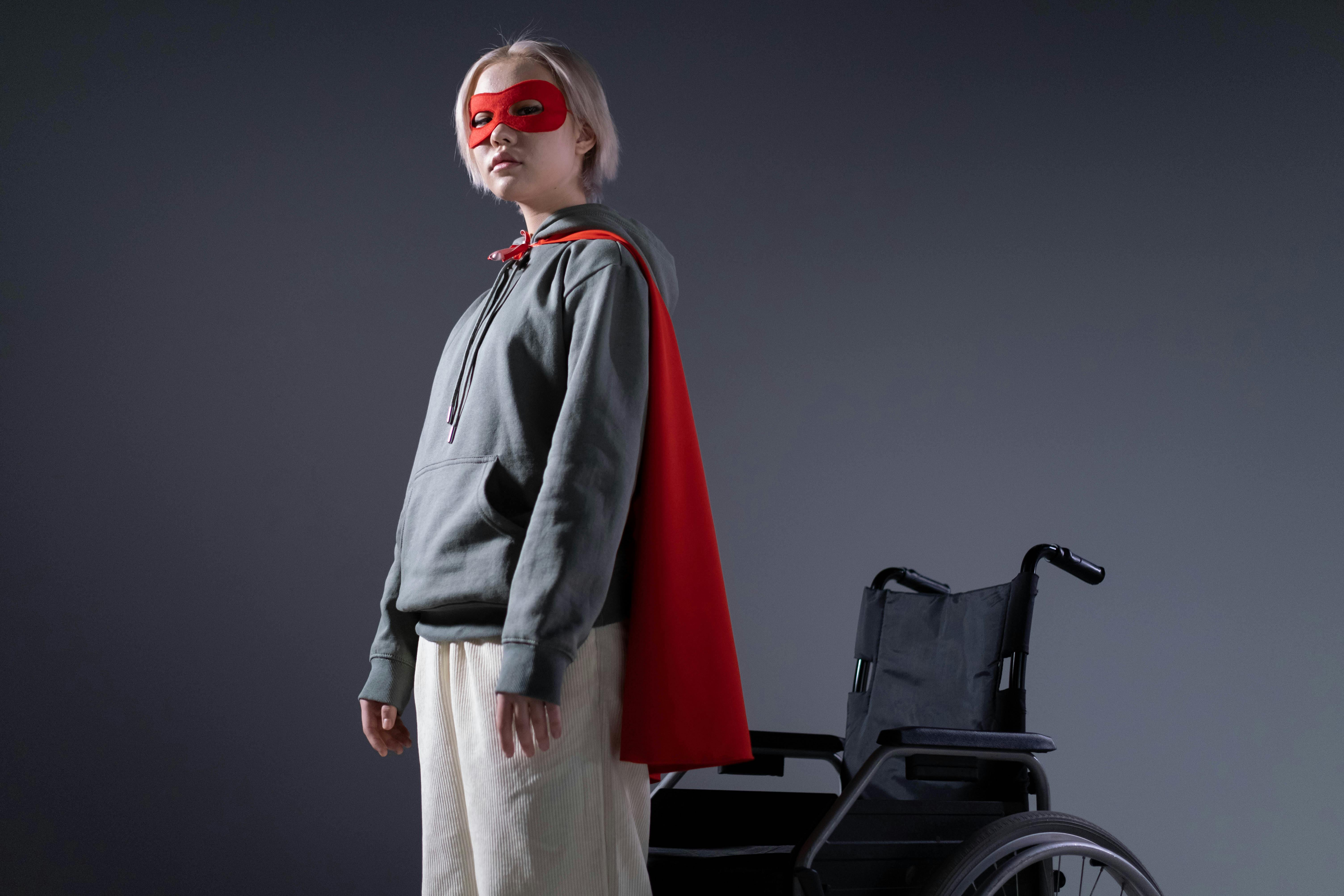 girl wearing a mask and cape standing beside a wheelchair