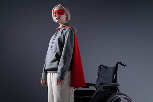 Girl Wearing a Mask and Cape Standing beside a Wheelchair