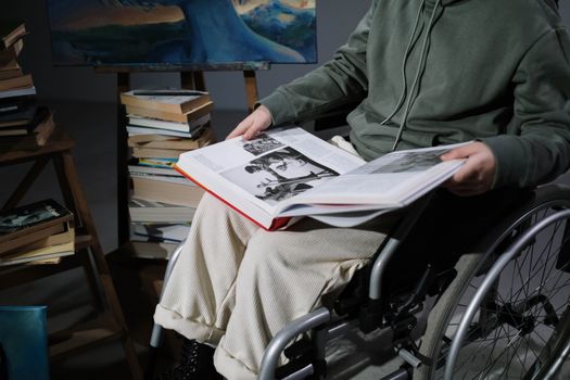 A person in a wheelchair reading a large book surrounded by stacks of books indoors.
