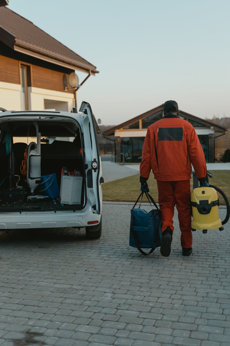 Man In Orange Work Clothes Standing Beside A Van Carrying Vaccum Cleaner And Bag