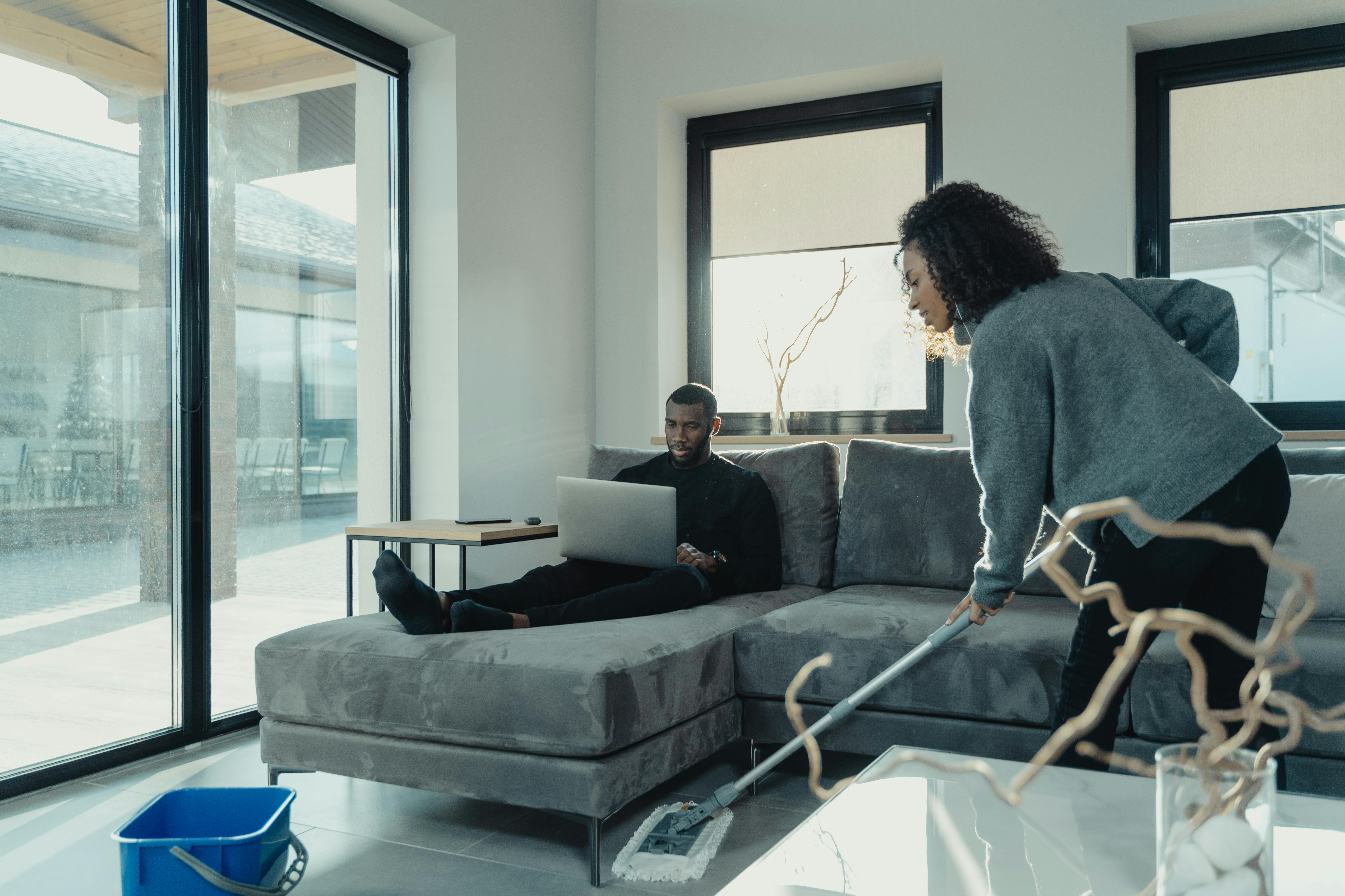 A couple in a modern living room, one using a laptop, the other cleaning.