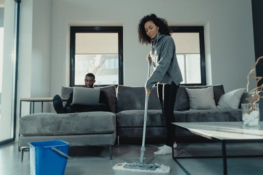 A woman mopping the floor while a man works on a laptop in a cozy modern living room.