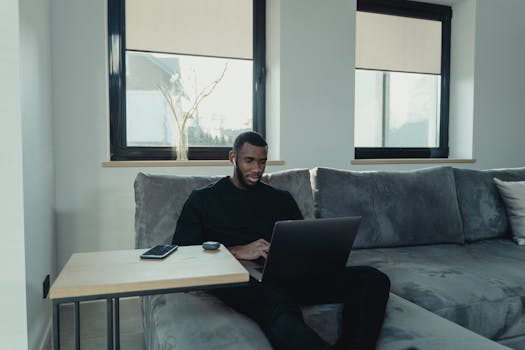 Man sitting on a sofa working on a laptop from home, with a cozy and modern interior setting.