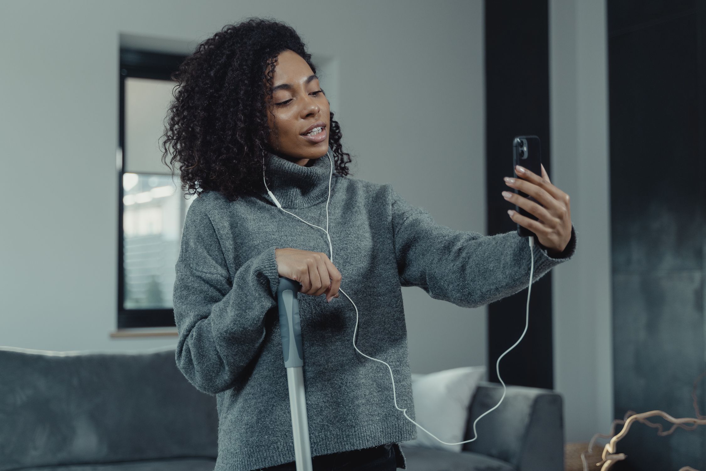 A woman enjoying a video call using earbuds as she cleans her living room with a mop.