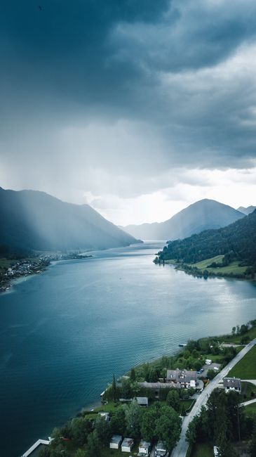 Capture of Weißensee, Austria showcasing dramatic clouds over serene lake surrounded by mountains and vivid landscape.