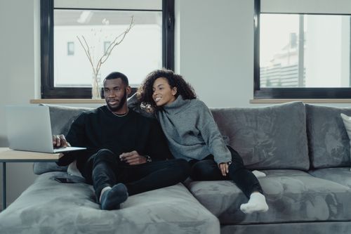 Free A joyful black couple enjoying time together on a sofa, browsing a laptop in a modern living room. Stock Photo