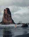 Back view of anonymous shirtless male jogging in wavy water of ocean with rough stony cliffs