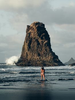 Back view of unrecognizable female in swimwear standing in waving ocean with rocky cliff and sandy beach under cloudy sky in daytime