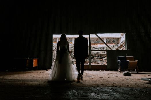 A bride and groom stand in a dark, deserted building, creating a dramatic silhouette.