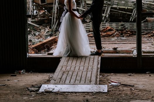 A couple holds hands in wedding attire inside an abandoned, decaying building.