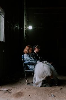 A contemplative couple in wedding attire sitting in an abandoned, dimly lit room.