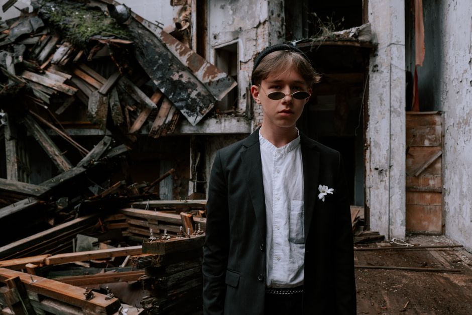 A young teen in stylish attire poses confidently in a derelict building setting.