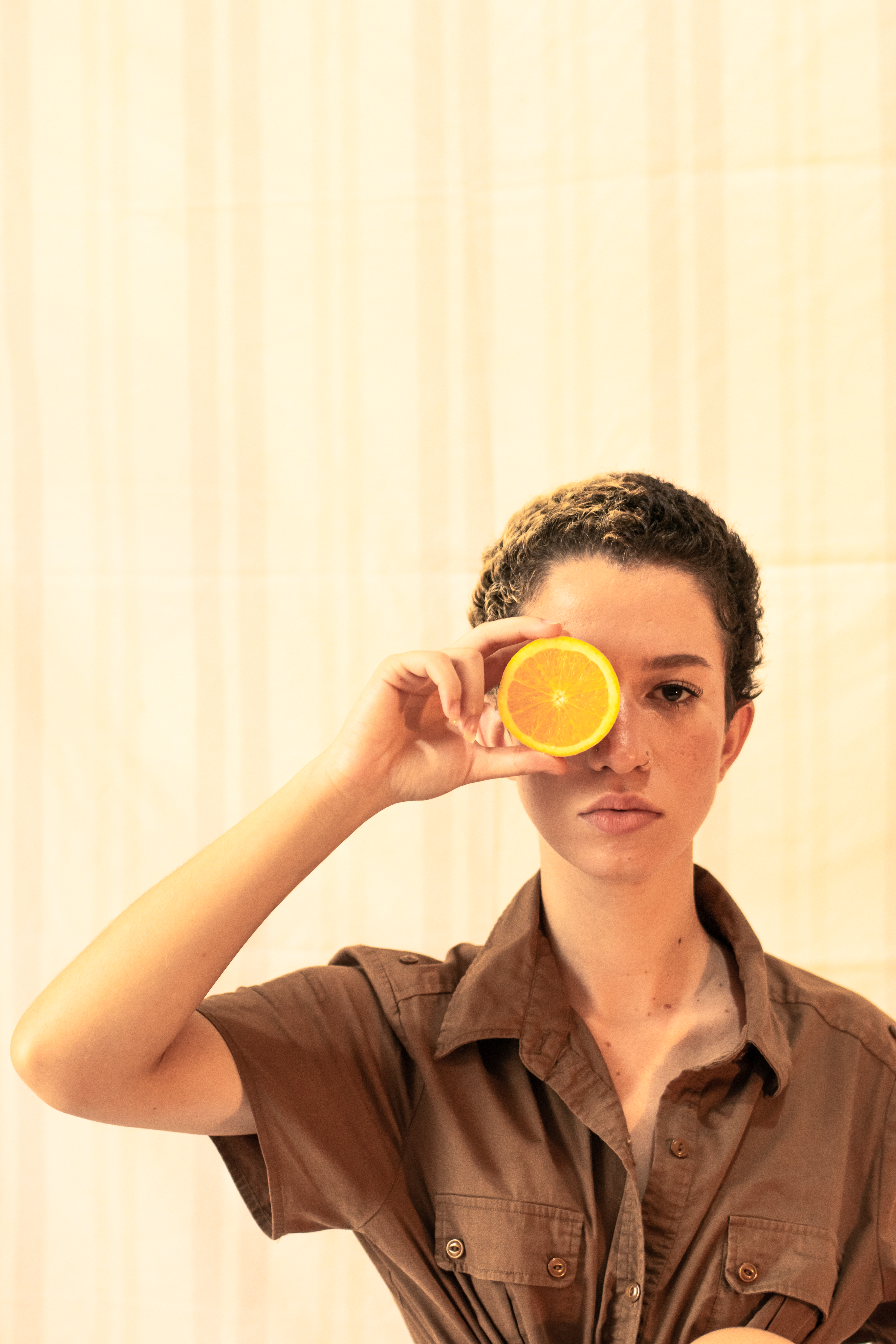 Portrait of a woman with short hair holding an orange slice over one eye, creating a playful pose.