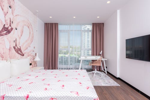 Comfortable room with wide bed under flamingos on walls against TV set and table near huge window