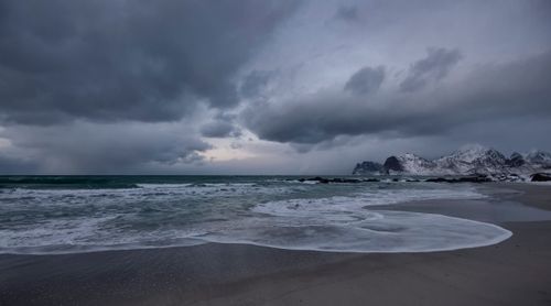 Free Dramatic coastal scenery with snow-capped mountains, dark clouds, and waves lapping the shore. Stock Photo