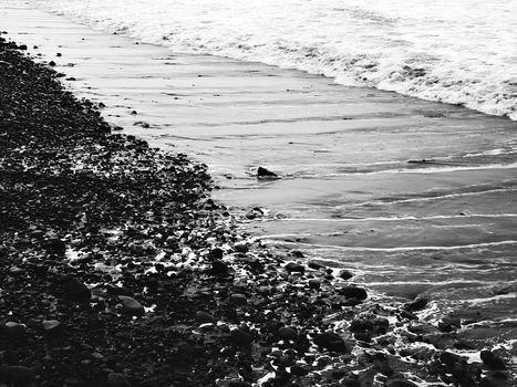 Black and white image of a pebble beach with waves crashing ashore, creating a serene shoreline scene.
