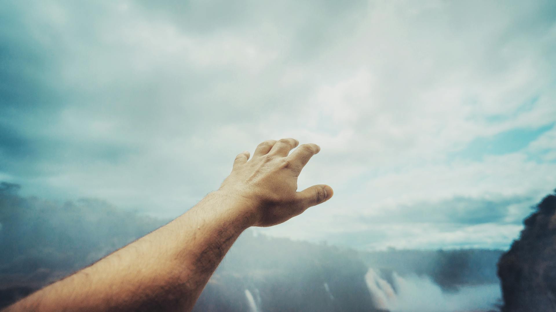 A hand reaching towards the cloudy sky at Iguazu Falls, depicting freedom and exploration.