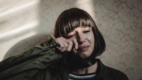 Unhappy female with modern haircut wiping tears while leaning on wall in room