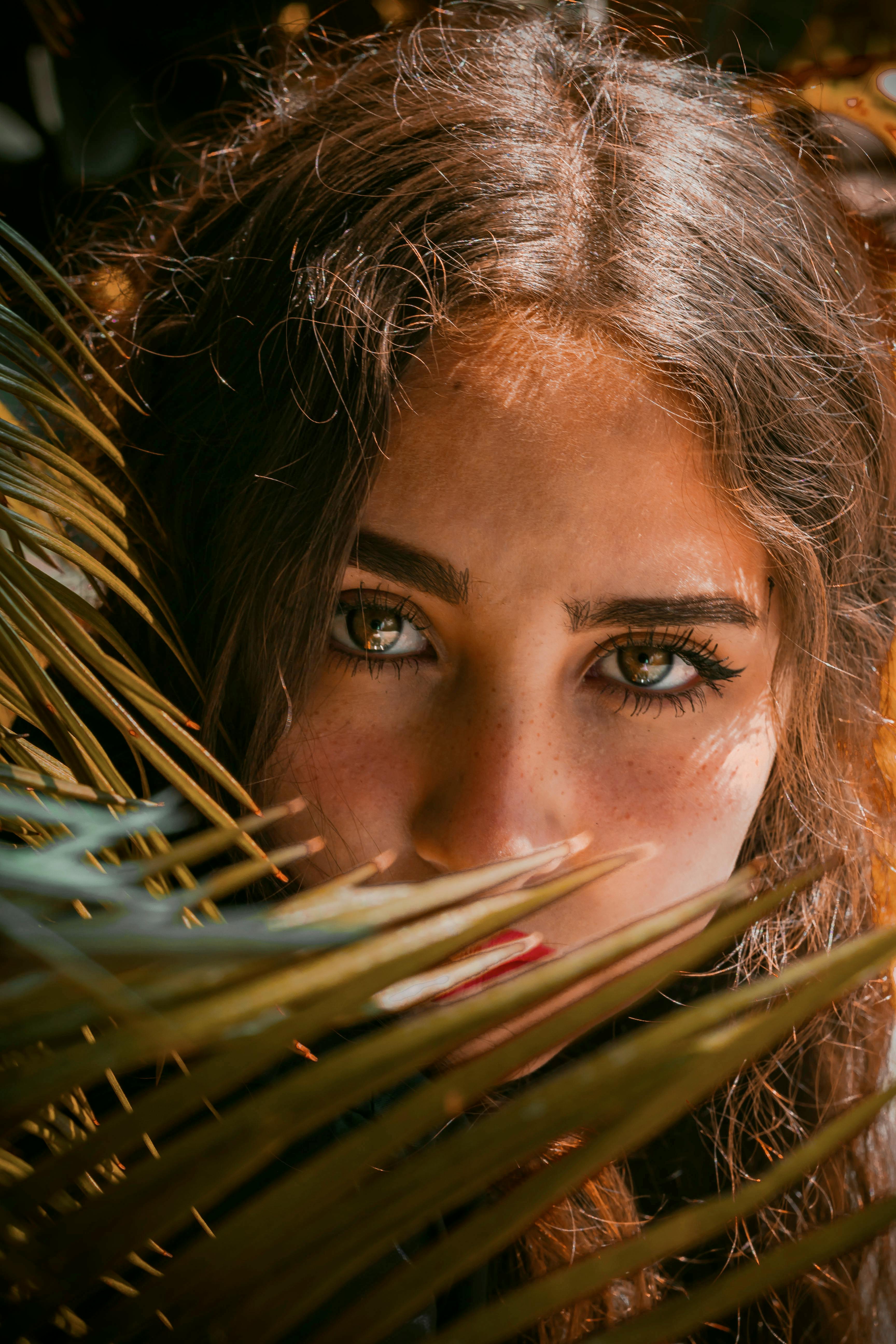 Close-up portrait of a woman peeking through palm leaves under daylight.