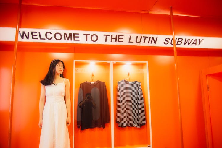 Girl In White Dress Standing Near Clothing On Hangers On Orange Walls