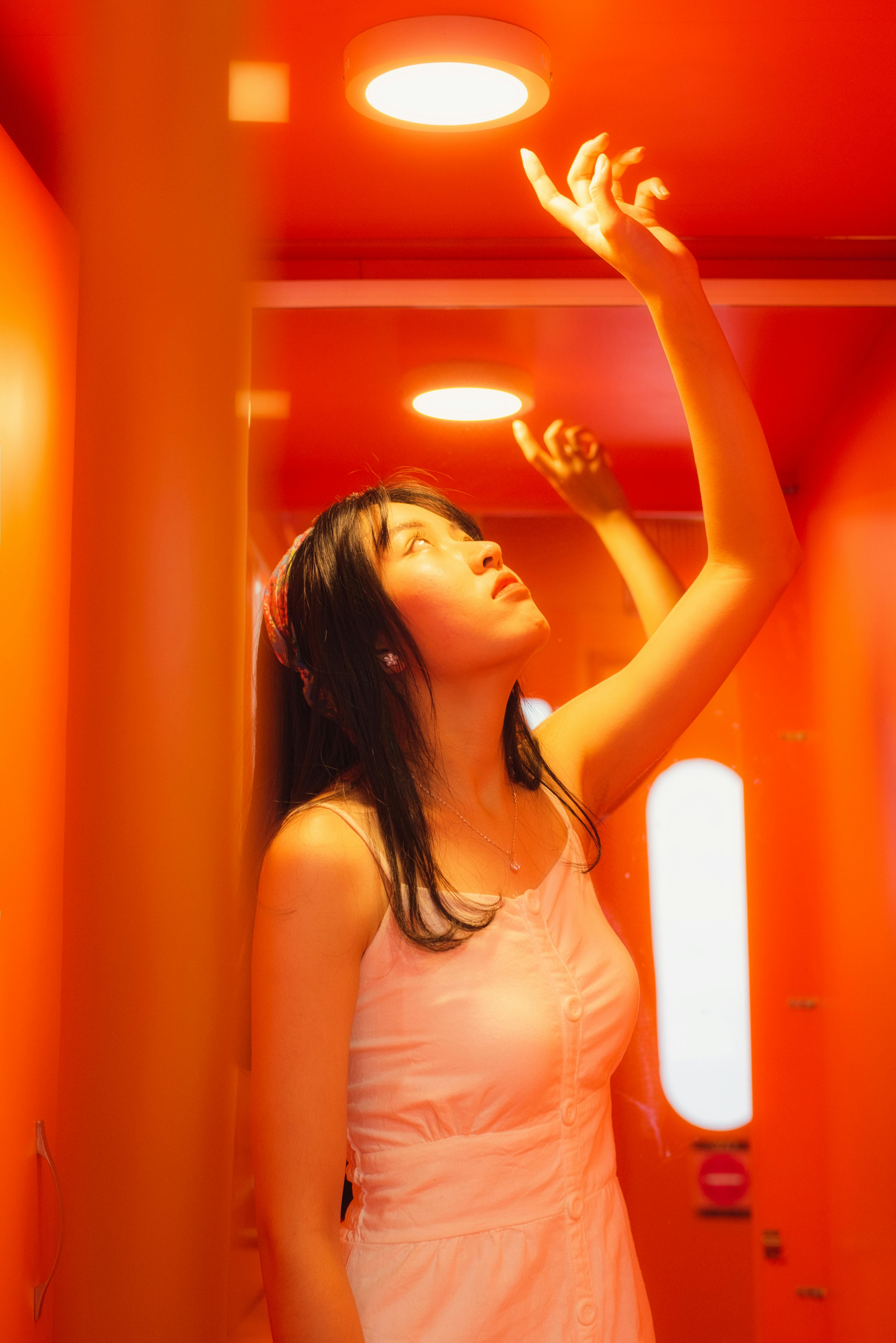 A woman in a white dress in an orange room, reaching towards ceiling lights that create a halo effect.