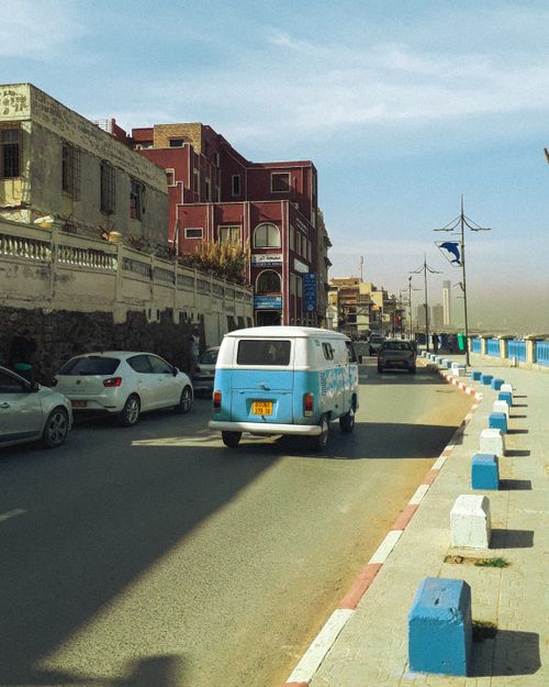 Free A classic van driving along a waterfront street with vintage buildings, creating a nostalgic scene. Stock Photo